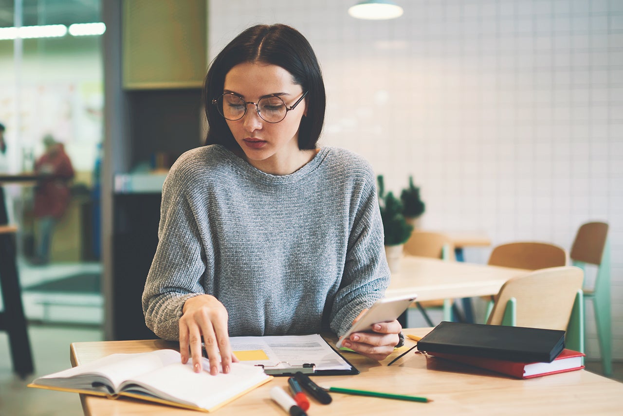 Skilled young female trainee of financial company making accounting report for restaurant income practicing due to advice of coach messaging via smartphone connected to free wireless internet