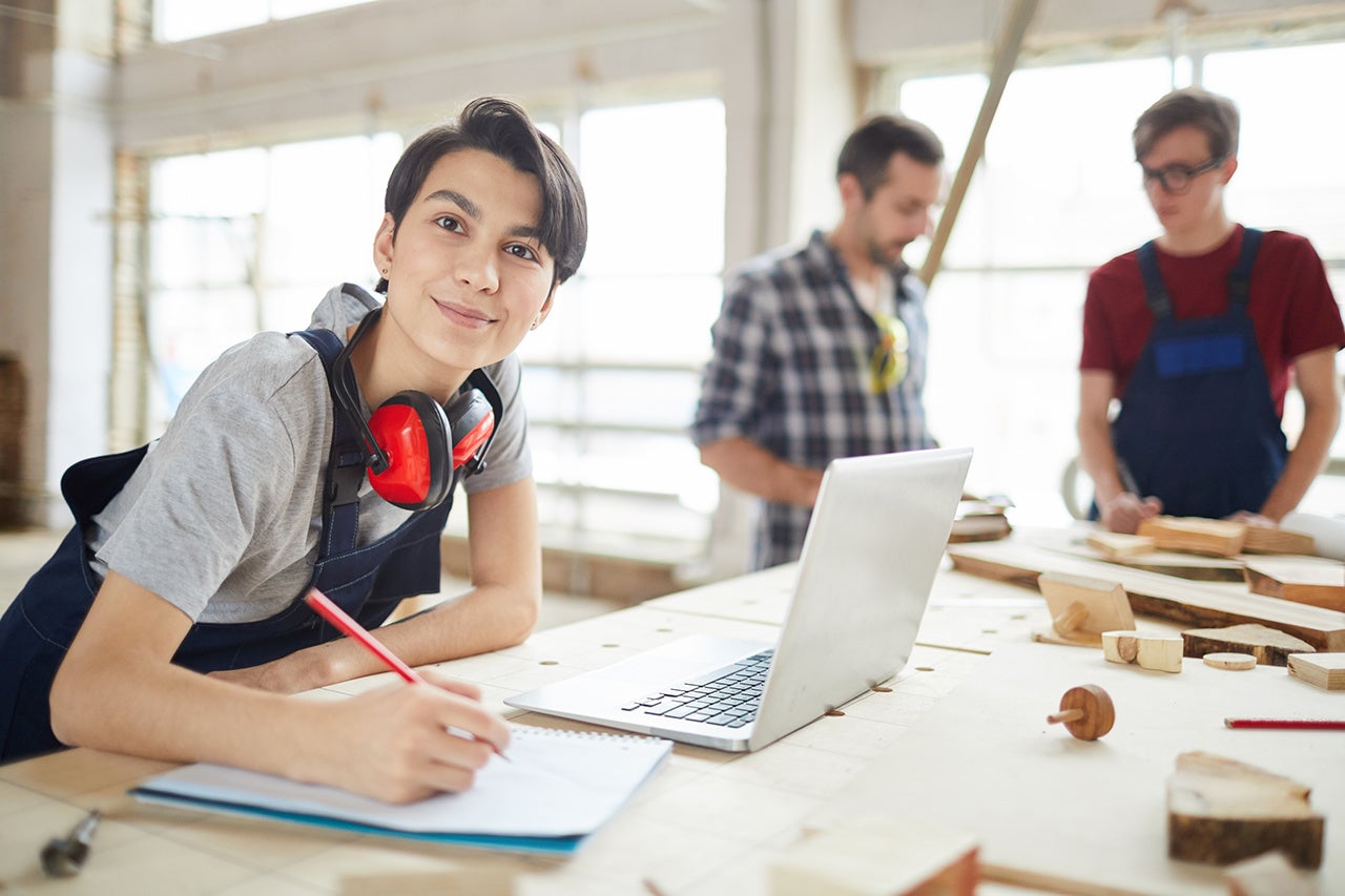 Smiling Young Woman Working in Carpenters Workshop, Portrait of modern carpenters working at woodworking factory, focus on young woman smiling at camera in foreground, copy space