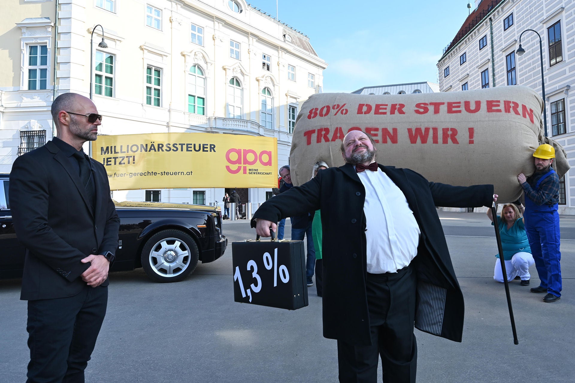 Foto: Im Vordergrund ein Millionär mit einem Koffer mit symbolischen 1,3% aufgedruckt, weil die Reichsten in Österreich mit vermögensbezogenen Steuern nur rund 3% zum Steueraufkommen beitragen. Im Hintergrund des Bildes sieht man einige Beschäftigte und "NormalverdienerInnen" - sie schultern die "Steuerlast" von 80%.