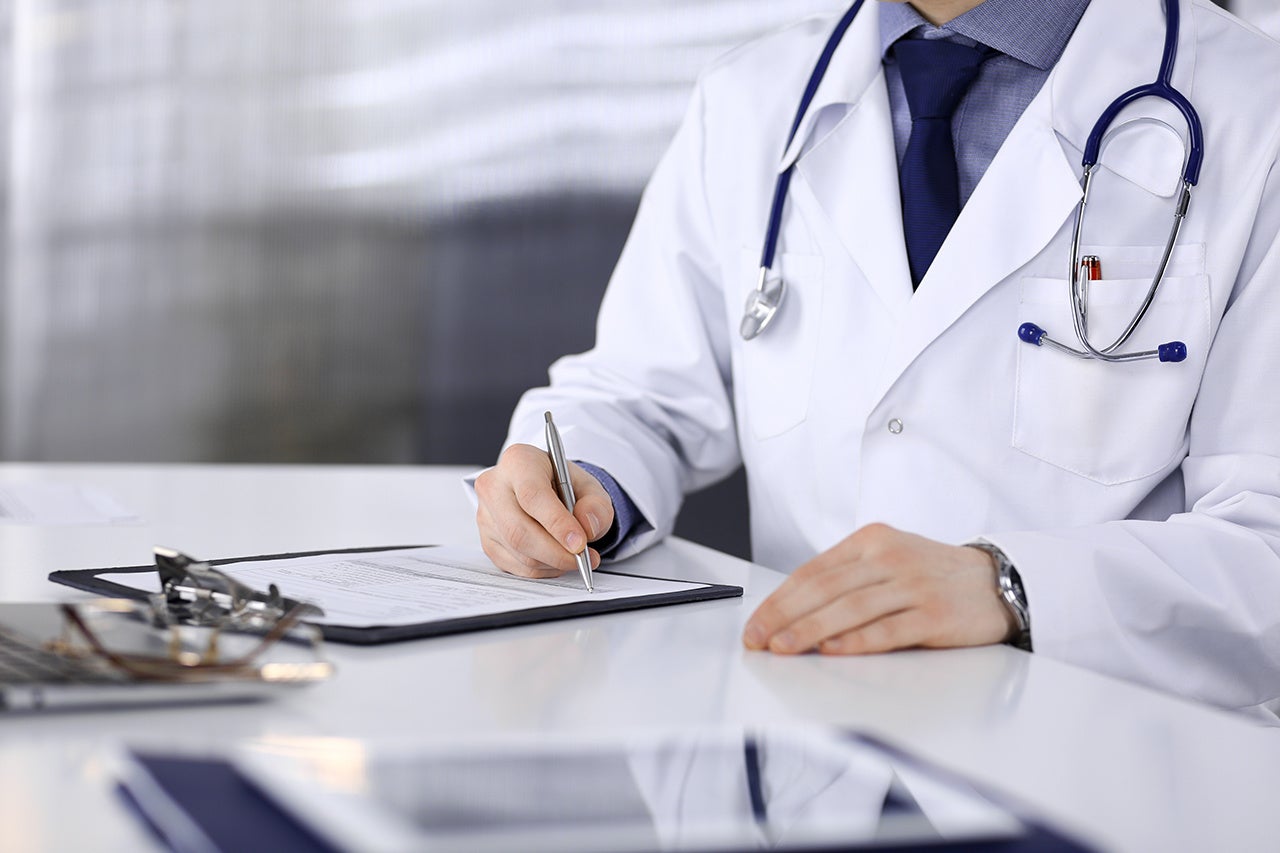 Unknown male doctor sitting and working with clipboard of medication history record in clinic at his working place, close-up. Young physician at work. Perfect medical service, medicine concept